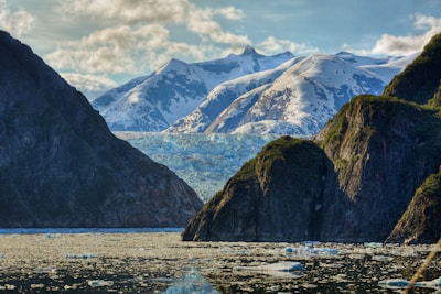 A picturesque landscape featuring snow-capped mountains in the background and rugged cliffs covered in green foliage in the foreground. A glacier spans across the scene, with chunks of ice floating on the calm water below. Fluffy clouds are scattered across a blue sky, enhancing the natural beauty of the serene environment.