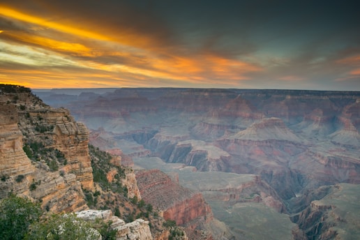 A vast and majestic canyon with intricate rock formations and cliffs, highlighted by a striking, colorful sunset sky. The landscape features deep valleys and rugged terrain while vegetation lines the canyon edges, creating a stunning contrast between the vibrant sky and the earthy tones of the rocks.