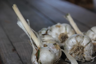 Several bulbs of garlic with long stems lie on a textured wooden surface. The garlic is in a natural, rustic state with some dried and peeling outer skins.