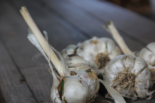 Several bulbs of garlic with long stems lie on a textured wooden surface. The garlic is in a natural, rustic state with some dried and peeling outer skins.