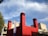 A modern architectural structure features three tall, rectangular red towers with louvered tops against a backdrop of a bright blue sky with wispy white clouds. The building has a contemporary design, combining concrete and red paneling. There is also a vertical banner with text, possibly advertising a show or exhibit.