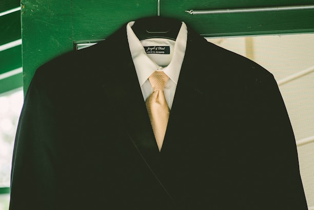 A black suit jacket hangs on a green door. The suit is complemented with a crisp white shirt and a beige tie. The jacket is displayed on a hanger, and the background features green wood and glass panes.