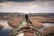 A person in a dark jacket walks along a large, rusty pipeline that stretches into the distance over a marshy landscape. The scene is set under an overcast sky, with sparse vegetation and bare trees surrounding the pipeline.