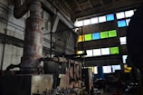 An industrial interior featuring large rusted machinery and pipes in a dimly lit room with high ceilings. The walls are concrete, and several rectangular windows are filled with colored panes in shades of blue, green, and yellow. The scene includes cluttered workspaces and scattered tools, creating an atmosphere of abandonment and decay.