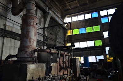 An industrial interior featuring large rusted machinery and pipes in a dimly lit room with high ceilings. The walls are concrete, and several rectangular windows are filled with colored panes in shades of blue, green, and yellow. The scene includes cluttered workspaces and scattered tools, creating an atmosphere of abandonment and decay.