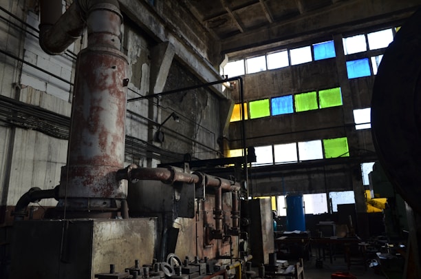 An industrial interior featuring large rusted machinery and pipes in a dimly lit room with high ceilings. The walls are concrete, and several rectangular windows are filled with colored panes in shades of blue, green, and yellow. The scene includes cluttered workspaces and scattered tools, creating an atmosphere of abandonment and decay.