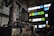 An industrial interior featuring large rusted machinery and pipes in a dimly lit room with high ceilings. The walls are concrete, and several rectangular windows are filled with colored panes in shades of blue, green, and yellow. The scene includes cluttered workspaces and scattered tools, creating an atmosphere of abandonment and decay.