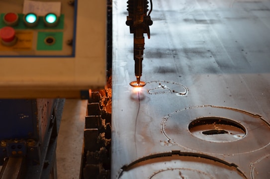 A machine is actively engaged in cutting or engraving a metallic surface with precision. The left side of the image shows a control panel with various buttons, one of which is red, and two indicator lights that are illuminated. The metallic surface has visible cut marks and circular shapes, indicating ongoing or completed cutting processes. The cutting tool emits a bright orange light as it makes contact with the metal.
