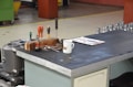 A workbench with various tools, a white mug, a paper with printed material, and metallic components. The tools include screwdrivers and other instruments placed upright on a block of wood. The surface is ribbed, and the backdrop suggests an industrial environment.