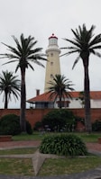 A tall lighthouse with a red top stands behind a row of palm trees. The structure is cream-colored with several small windows along its height. In front of it, there is a brick building with a red tile roof. The area is surrounded by well-maintained greenery, including trimmed bushes and grass, and a circular path with a star-shaped decoration.