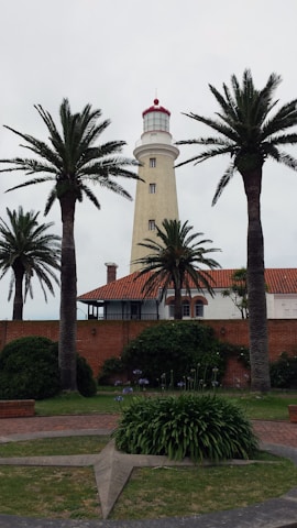 A tall lighthouse with a red top stands behind a row of palm trees. The structure is cream-colored with several small windows along its height. In front of it, there is a brick building with a red tile roof. The area is surrounded by well-maintained greenery, including trimmed bushes and grass, and a circular path with a star-shaped decoration.