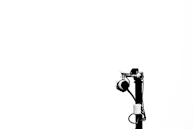 A black and white image featuring a surveillance camera mounted on a tall pole. The background is a plain white, enhancing the stark contrast and minimalist composition. The camera is positioned to face away from the viewer, with various wires and components visible.