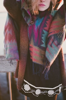 A person is wearing a colorful, patterned poncho and a black outfit with a decorative hem featuring white floral designs. The individual is standing outside, with part of their face and blonde hair visible, and appears to be by a wooden structure.