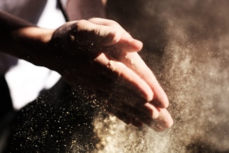 Hands clapping with flour or powder particles spreading in the air, creating a dynamic and dramatic visual effect. The scene is illuminated by soft lighting, highlighting the fine details of the particles.