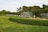 Fountains gracefully spraying water are set against a meticulously maintained green lawn, bordered by well-manicured hedges. A backdrop of lush, dense trees frames the scene, with a clear sky above.