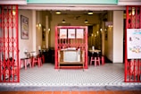 A retro-style café interior with red metal gates and vintage decorations. The center features a vintage television surrounded by wooden tables and red chairs. The walls display framed art and menus, while pendant lights hang from the ceiling. The floor is patterned with small black and white tiles.