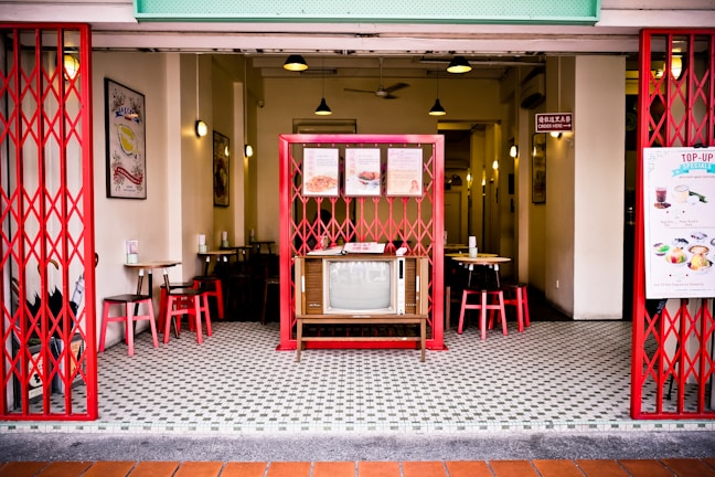 A retro-style café interior with red metal gates and vintage decorations. The center features a vintage television surrounded by wooden tables and red chairs. The walls display framed art and menus, while pendant lights hang from the ceiling. The floor is patterned with small black and white tiles.