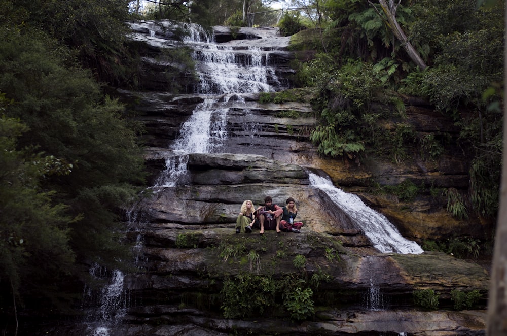 three person sitting on rock formation in waterfalls surrounded with trees at daytime