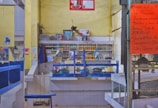 A small food stall or restaurant with a kitchen setup, featuring a counter with various cooking pots and utensils. The wall shelves behind the counter hold condiments and containers. The walls are painted yellow, and there are various posters and photos displayed. A large orange menu is posted on a chain-link fence to the right, listing various food offerings in Spanish.