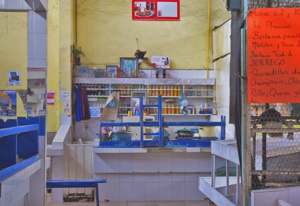 A small food stall or restaurant with a kitchen setup, featuring a counter with various cooking pots and utensils. The wall shelves behind the counter hold condiments and containers. The walls are painted yellow, and there are various posters and photos displayed. A large orange menu is posted on a chain-link fence to the right, listing various food offerings in Spanish.