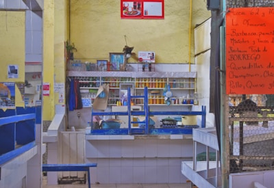 A small food stall or restaurant with a kitchen setup, featuring a counter with various cooking pots and utensils. The wall shelves behind the counter hold condiments and containers. The walls are painted yellow, and there are various posters and photos displayed. A large orange menu is posted on a chain-link fence to the right, listing various food offerings in Spanish.