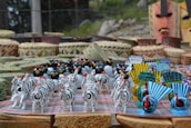 A collection of colorful handcrafted figurines including zebras, birds, and masks is neatly arranged on a table. Baskets and woven items serve as a backdrop, enhancing the artisanal display.