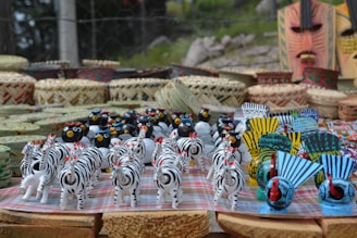 A collection of colorful handcrafted figurines including zebras, birds, and masks is neatly arranged on a table. Baskets and woven items serve as a backdrop, enhancing the artisanal display.