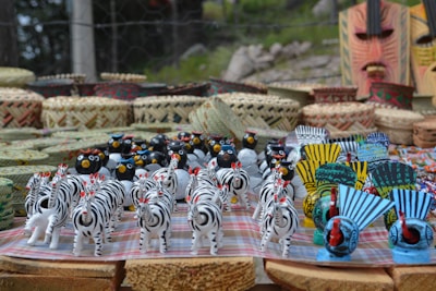 A collection of colorful handcrafted figurines including zebras, birds, and masks is neatly arranged on a table. Baskets and woven items serve as a backdrop, enhancing the artisanal display.