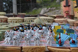 A collection of colorful handcrafted figurines including zebras, birds, and masks is neatly arranged on a table. Baskets and woven items serve as a backdrop, enhancing the artisanal display.