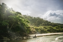 A lush, green tropical landscape with dense foliage and a serene beach setting. Sunlight filters through the trees, creating a tranquil atmosphere. A person appears to be sitting on the rocks near the shoreline, adding a sense of calm and relaxation to the scene.