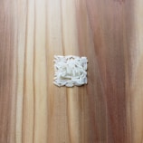 A small pile of uncooked white rice grains is arranged in a square shape on a wooden surface. The wooden surface has a variety of light and dark brown tones, showing a natural grain pattern.