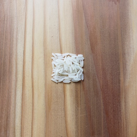 A small pile of uncooked white rice grains is arranged in a square shape on a wooden surface. The wooden surface has a variety of light and dark brown tones, showing a natural grain pattern.