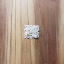 A small pile of uncooked white rice grains is arranged in a square shape on a wooden surface. The wooden surface has a variety of light and dark brown tones, showing a natural grain pattern.