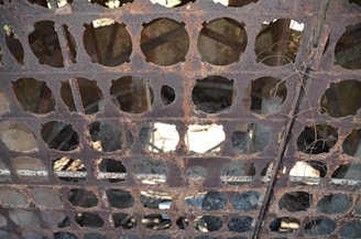 A rusted metal grate with circular cutouts is elevated, casting intricate shadows beneath it. The background shows a partially visible, weathered industrial structure with light filtering through gaps.