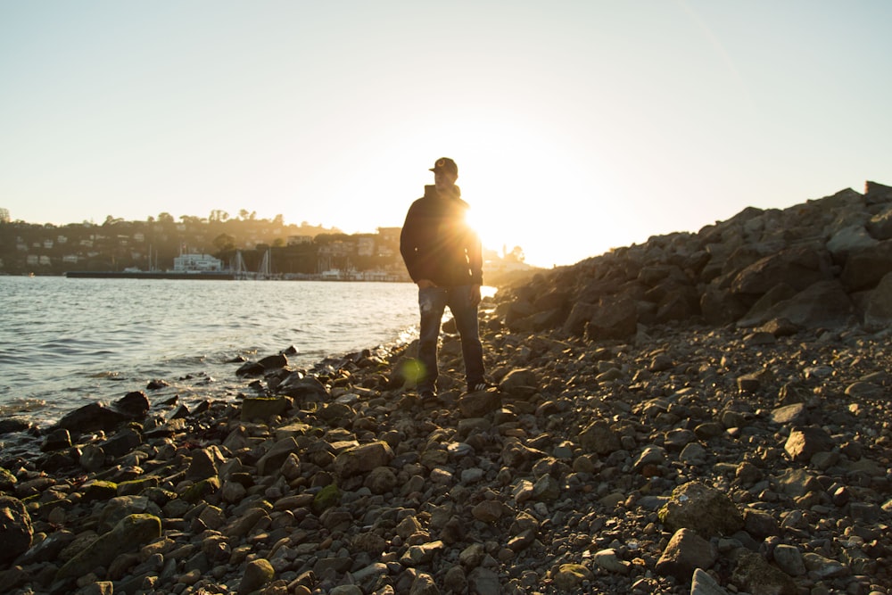 man wearing black jacket standing on stones near body of water under blue sky during golden hour