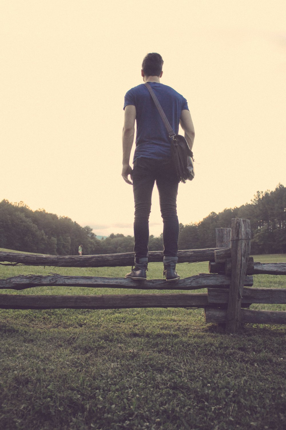 man in blue t-shirt with black denim jeans and pair of gray high-top sneakers standing brown wooden fence during daytime