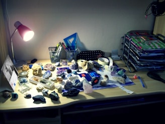 A wooden desk holds an assortment of rocks and minerals scattered across its surface. A bright desk lamp illuminates the area, casting shadows over various tools and stationery. In the background, mesh pencil holders contain pens and markers, while an organizer on the right side holds stacked colorful files and papers. A blue ruler and black comb are also visible.