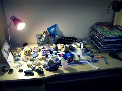 A wooden desk holds an assortment of rocks and minerals scattered across its surface. A bright desk lamp illuminates the area, casting shadows over various tools and stationery. In the background, mesh pencil holders contain pens and markers, while an organizer on the right side holds stacked colorful files and papers. A blue ruler and black comb are also visible.