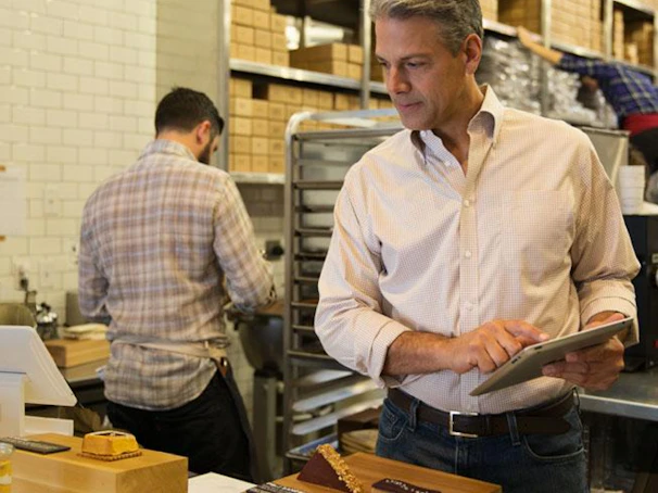 A café owner happily reviewing online orders on a tablet in a cozy shop setting.
