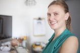 A person with a cheerful smile is seated at a desk in an office setting. They are wearing a sleeveless teal top and have earrings. In the background, there are office items like a computer monitor, papers, and blurred objects.
