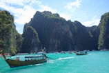 A picturesque landscape featuring a serene turquoise sea surrounded by towering, lush green cliffs. Several traditional long-tail boats with tourists can be seen on the water, set against a backdrop of bright blue skies dotted with scattered clouds.