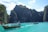 A picturesque landscape featuring a serene turquoise sea surrounded by towering, lush green cliffs. Several traditional long-tail boats with tourists can be seen on the water, set against a backdrop of bright blue skies dotted with scattered clouds.