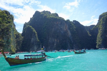 A picturesque landscape featuring a serene turquoise sea surrounded by towering, lush green cliffs. Several traditional long-tail boats with tourists can be seen on the water, set against a backdrop of bright blue skies dotted with scattered clouds.