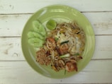 A plate of pad thai with shrimp, peanuts, bean sprouts, and an egg, garnished with sliced cucumber and a lime wedge, presented on a light green plate on a rustic wooden table.