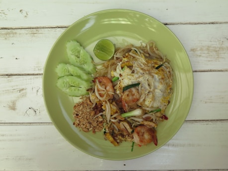 A plate of pad thai with shrimp, peanuts, bean sprouts, and an egg, garnished with sliced cucumber and a lime wedge, presented on a light green plate on a rustic wooden table.