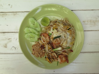 A plate of pad thai with shrimp, peanuts, bean sprouts, and an egg, garnished with sliced cucumber and a lime wedge, presented on a light green plate on a rustic wooden table.