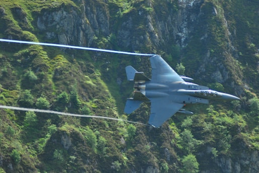 A fighter jet flies at high speed through a lush, green mountainous landscape. The jet's sleek design is accentuated by the contrast against the rugged terrain, with trails of condensation visible behind the wings.