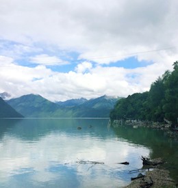 A tranquil alpine lake surrounded by mountains under a partly cloudy sky reflects the scenery on its still surface. Lush green trees line the shoreline on the right, with scattered driftwood and stones along the water's edge.