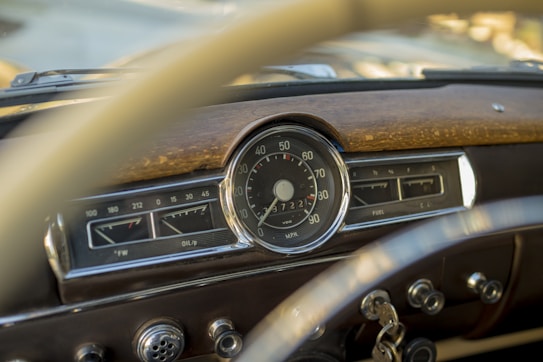 A close-up view of a vintage car dashboard featuring a speedometer, fuel gauge, and oil pressure gauge. The display includes chrome details and a wooden panel accent. Classic car keys are hanging from the ignition.