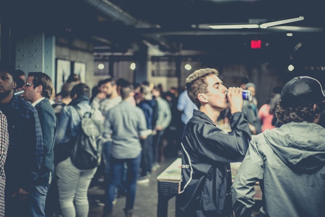 A crowded indoor gathering with people socializing and some holding drinks. The setting appears to be a casual and relaxed environment with dim lighting and a mix of individuals in conversation.